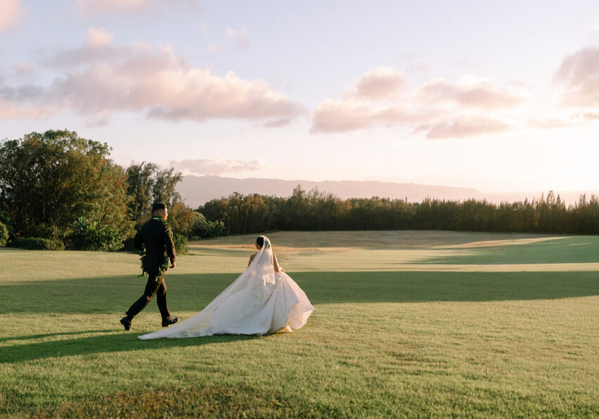 A bride and groom walk across a grassy field under a partly cloudy sky during sunset.