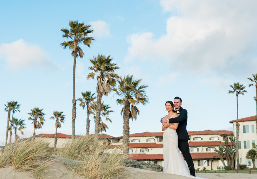 A couple stands on a sandy beach with palm trees and buildings in the background under a partly cloudy sky.