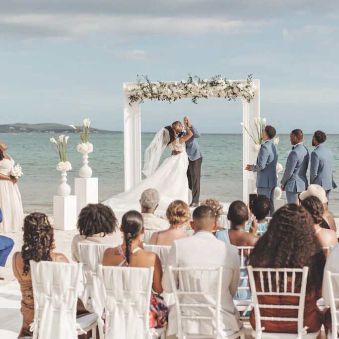 A bride and groom kiss under a floral arch on a beach as wedding guests seated outdoors look on.