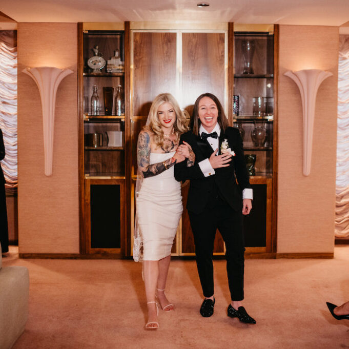 A couple in wedding attire enters a room, smiling. The person on the left wears a dress; the one on the right wears a tuxedo. An attendee applauds in the background.