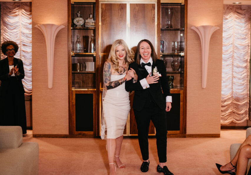 A couple in wedding attire enters a room, smiling. The person on the left wears a dress; the one on the right wears a tuxedo. An attendee applauds in the background.