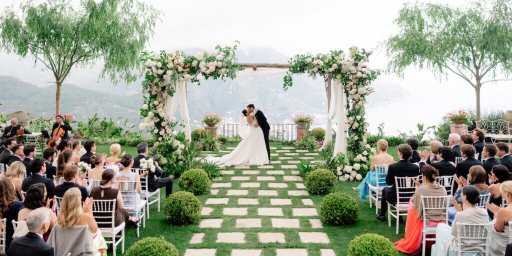 Bride and groom kiss under a flower-adorned arch at an outdoor wedding ceremony, with guests seated on either side of a stone path surrounded by greenery.
