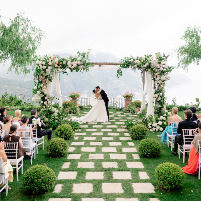 Bride and groom kiss under a flower-adorned arch at an outdoor wedding ceremony, with guests seated on either side of a stone path surrounded by greenery.
