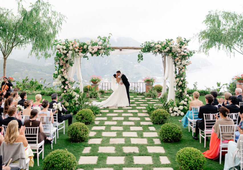 Bride and groom kiss under a flower-adorned arch at an outdoor wedding ceremony, with guests seated on either side of a stone path surrounded by greenery.
