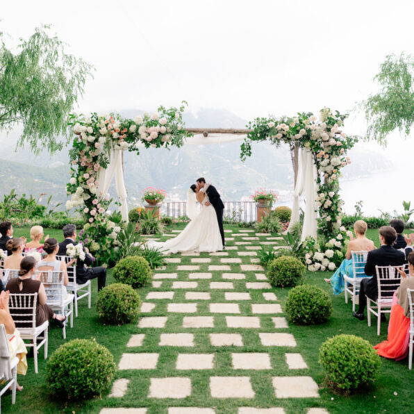 Bride and groom kiss under a flower-adorned arch at an outdoor wedding ceremony, with guests seated on either side of a stone path surrounded by greenery.