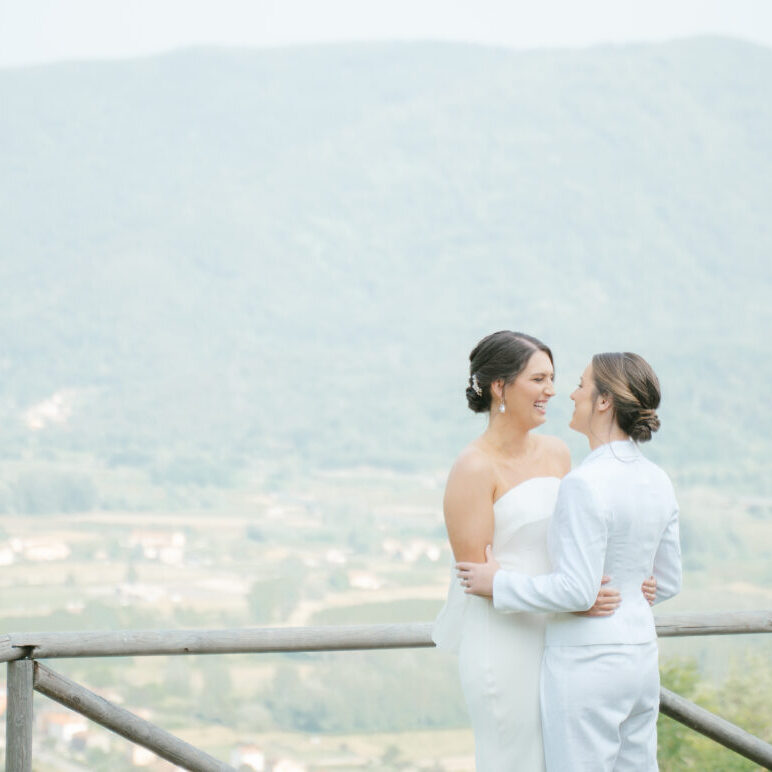 Two women in wedding attire stand close together, smiling at each other on a wooden terrace with a scenic, mountainous landscape in the background.