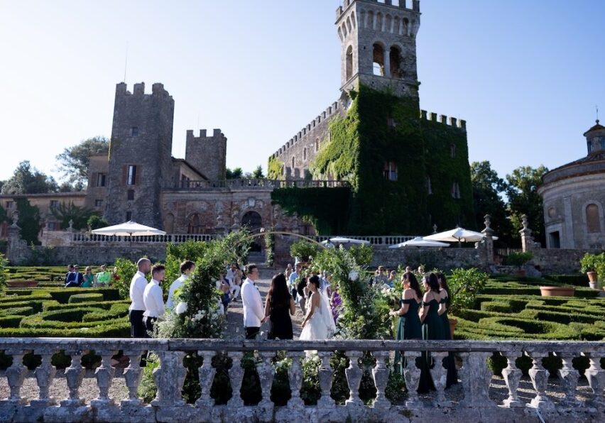 A group of people are gathered outdoors at a wedding ceremony, standing in front of a large, ivy-covered castle with turrets. The setting includes well-manicured gardens and hedges, with clear blue skies in the background. Guests are wearing formal attire.