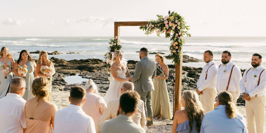 A couple stands under a floral arch exchanging vows during a beach wedding ceremony, with guests seated and an officiant present.