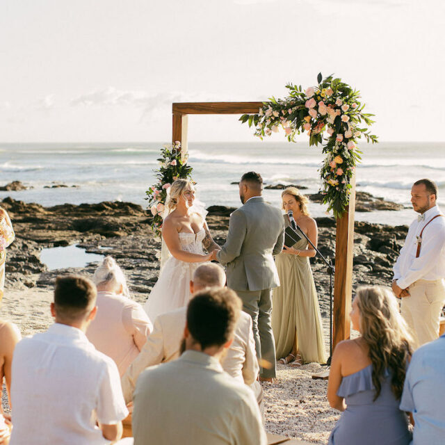 A couple stands under a floral arch exchanging vows during a beach wedding ceremony, with guests seated and an officiant present.