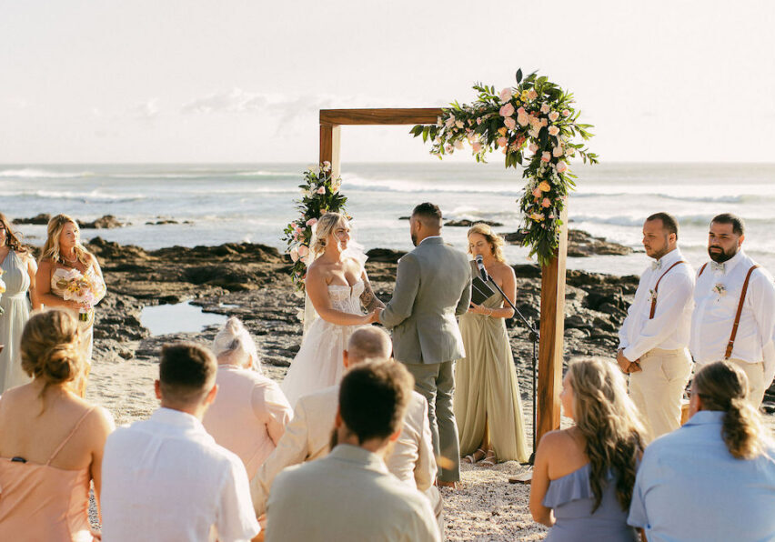 A couple stands under a floral arch exchanging vows during a beach wedding ceremony, with guests seated and an officiant present.