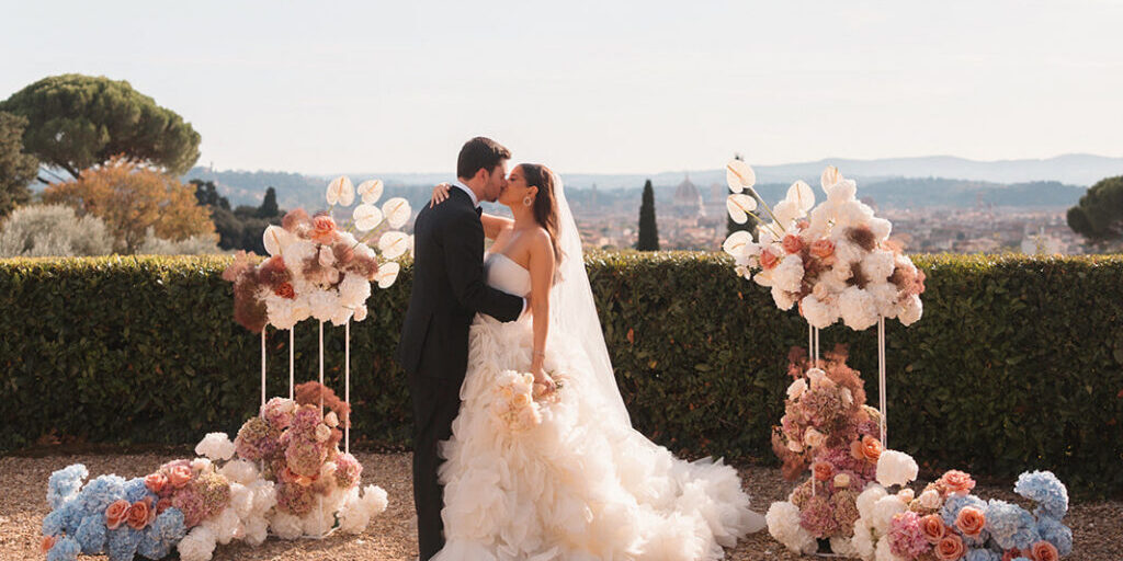 A bride and groom kiss outdoors between two floral arrangements, with a scenic landscape and city in the background.