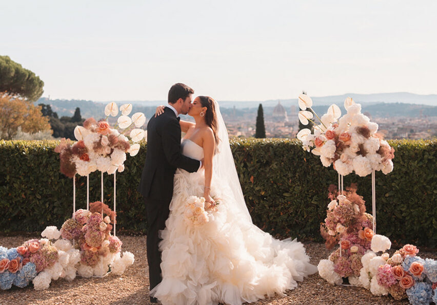A bride and groom kiss outdoors between two floral arrangements, with a scenic landscape and city in the background.