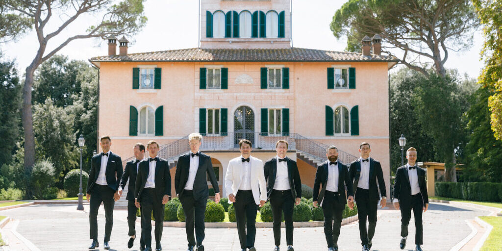 A group of men in formal suits, one in a white jacket, walk in front of a large, elegant villa with green shutters and arched windows.
