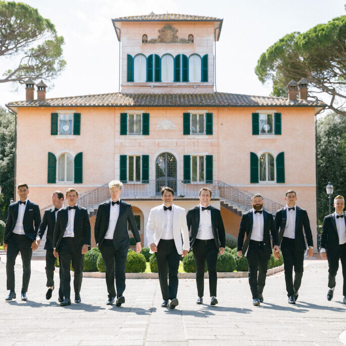 A group of men in formal suits, one in a white jacket, walk in front of a large, elegant villa with green shutters and arched windows.