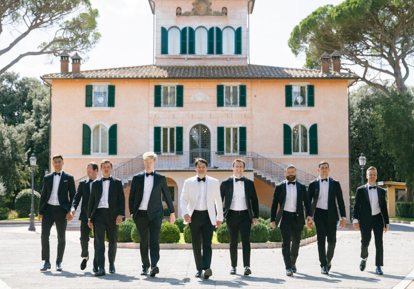 A group of men in formal suits, one in a white jacket, walk in front of a large, elegant villa with green shutters and arched windows.