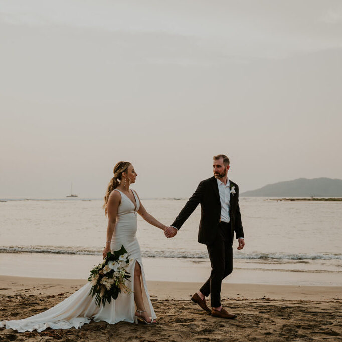 A couple in formal attire walks hand in hand on a sandy beach with the sea and distant mountains in the background. The bride holds a bouquet in her left hand.