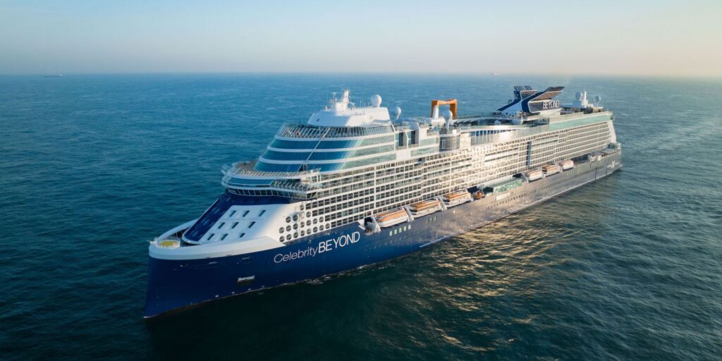 A large blue and white cruise ship sails on calm ocean waters under a clear sky.