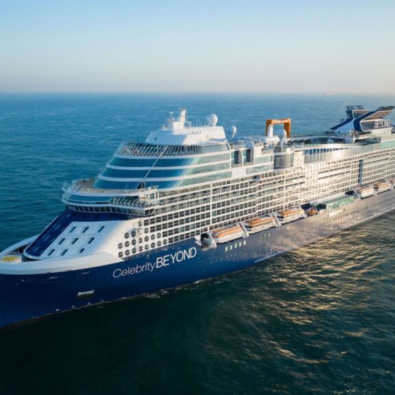 A large blue and white cruise ship sails on calm ocean waters under a clear sky.
