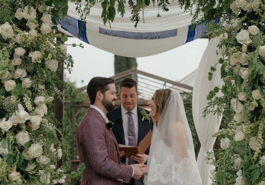 A bride and groom stand under a floral arch, holding hands and smiling, while an officiant reads from a book during an outdoor wedding ceremony.