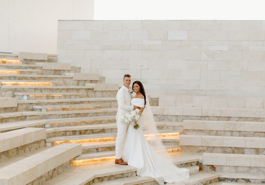 A bride and groom pose together on illuminated stone steps in front of a white stone wall. The bride holds a bouquet and wears an off-the-shoulder dress with a train and veil.