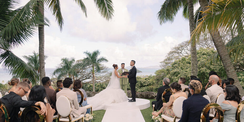 A bride and groom stand at an outdoor altar surrounded by guests, with palm trees and a view of the ocean in the background.
