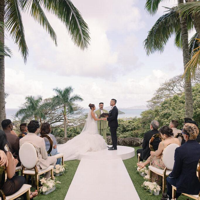A bride and groom stand at an outdoor altar surrounded by guests, with palm trees and a view of the ocean in the background.