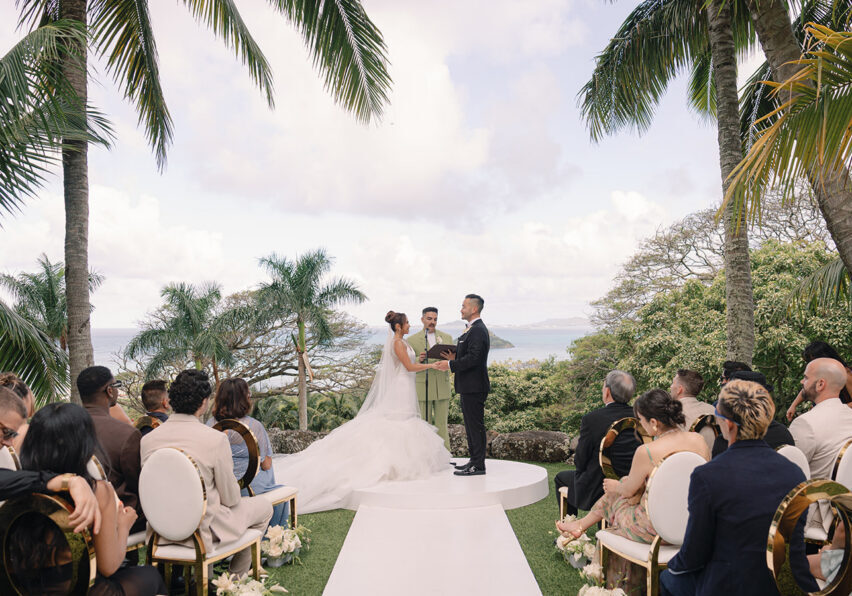 A bride and groom stand at an outdoor altar surrounded by guests, with palm trees and a view of the ocean in the background.
