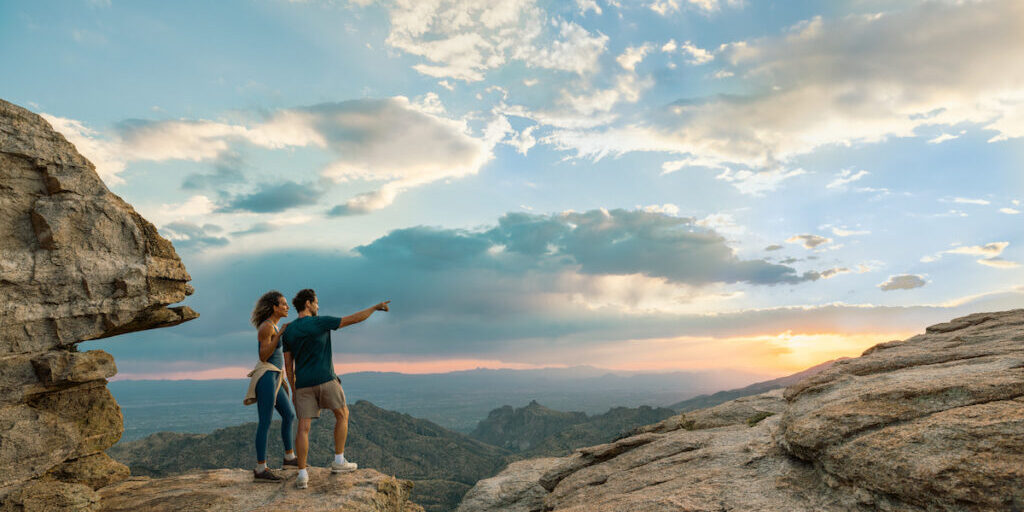 Two people stand on a rocky cliff at sunset, with one person pointing toward the horizon; mountains and a dramatic sky are visible in the background.