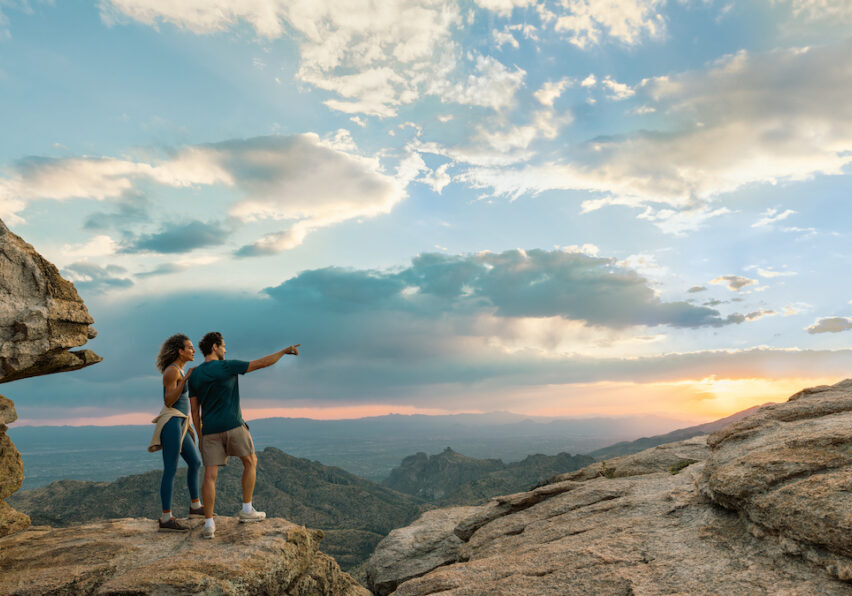 Two people stand on a rocky cliff at sunset, with one person pointing toward the horizon; mountains and a dramatic sky are visible in the background.