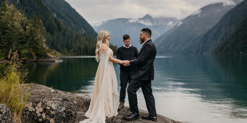 A couple is getting married outdoors by a lake, surrounded by mountains, while an officiant conducts the ceremony.