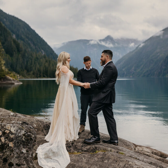 A couple is getting married outdoors by a lake, surrounded by mountains, while an officiant conducts the ceremony.