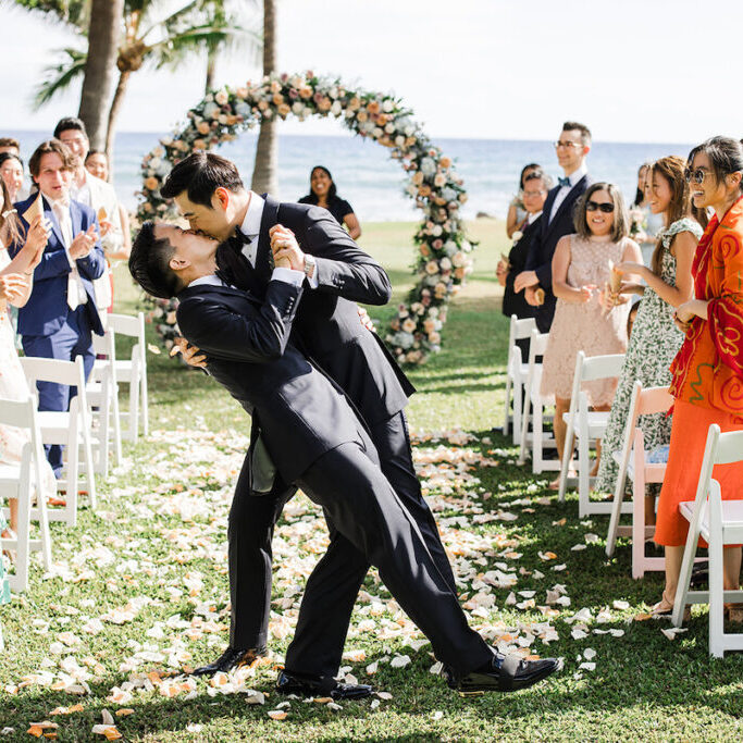 groom dips and kisses his groom with wedding guests clapping to the side and circle floral arch and ocean in background