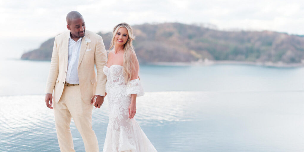 A couple in wedding attire stands by an infinity pool, smiling at each other. The background features a calm seascape and distant island.