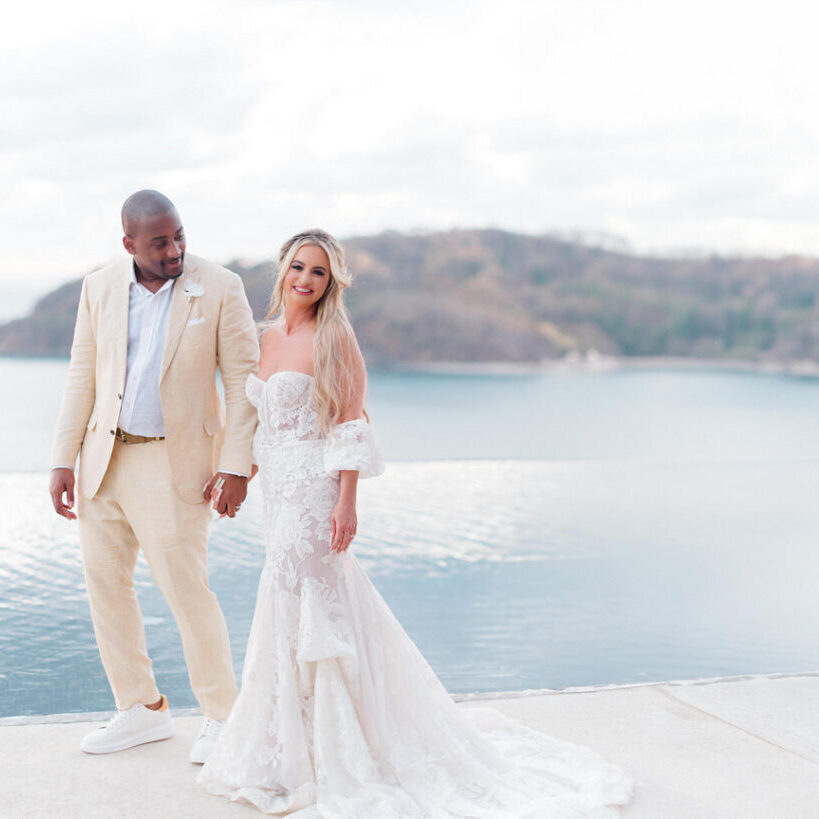 A couple in wedding attire stands by an infinity pool, smiling at each other. The background features a calm seascape and distant island.