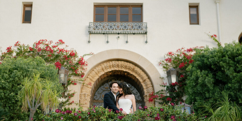 A couple dressed formally stands close together on a balcony in front of a large arched doorway, surrounded by flowering plants and greenery.