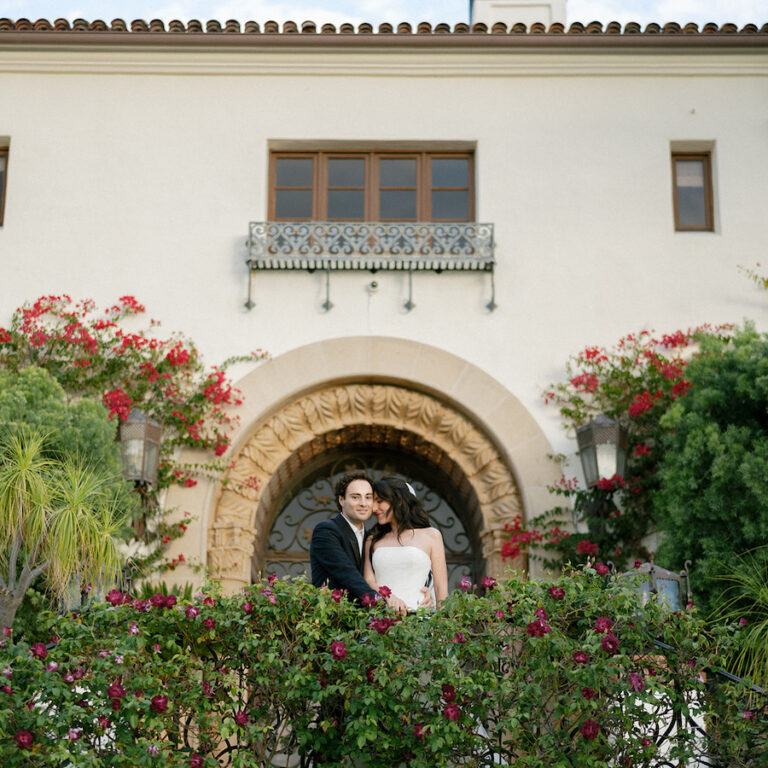 A couple dressed formally stands close together on a balcony in front of a large arched doorway, surrounded by flowering plants and greenery.