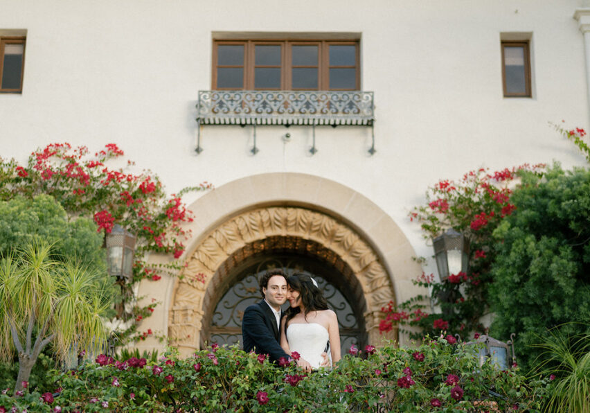 A couple dressed formally stands close together on a balcony in front of a large arched doorway, surrounded by flowering plants and greenery.