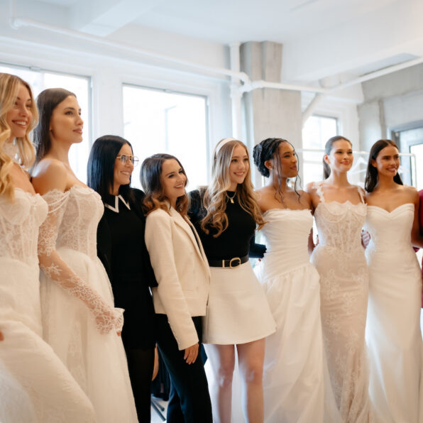 A group of women, some in wedding dresses and others in business attire, stand together and pose for a photo in a bright room.