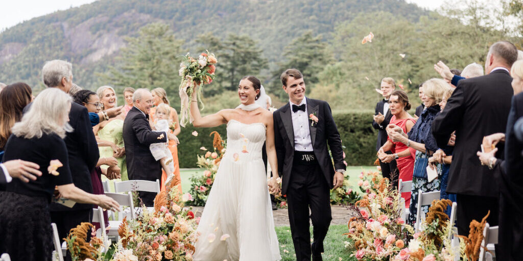 A bride and groom walk down an outdoor aisle lined with flowers, smiling and holding hands, as guests celebrate around them with flower petals. Mountains and trees are visible in the background.