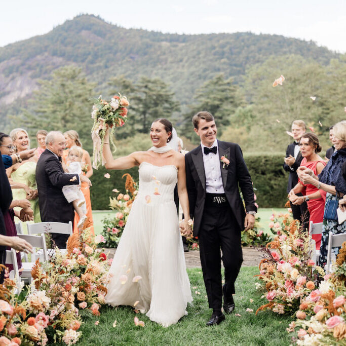 A bride and groom walk down an outdoor aisle lined with flowers, smiling and holding hands, as guests celebrate around them with flower petals. Mountains and trees are visible in the background.