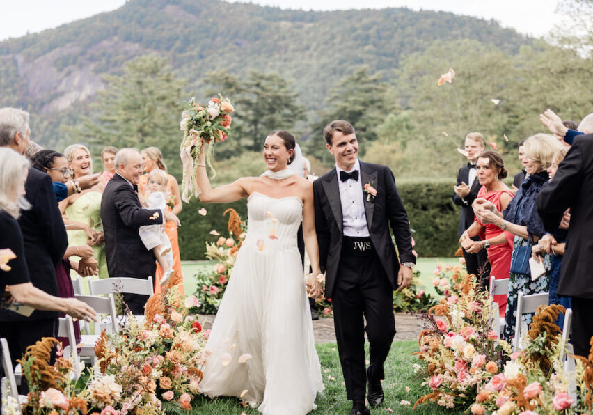 A bride and groom walk down an outdoor aisle lined with flowers, smiling and holding hands, as guests celebrate around them with flower petals. Mountains and trees are visible in the background.