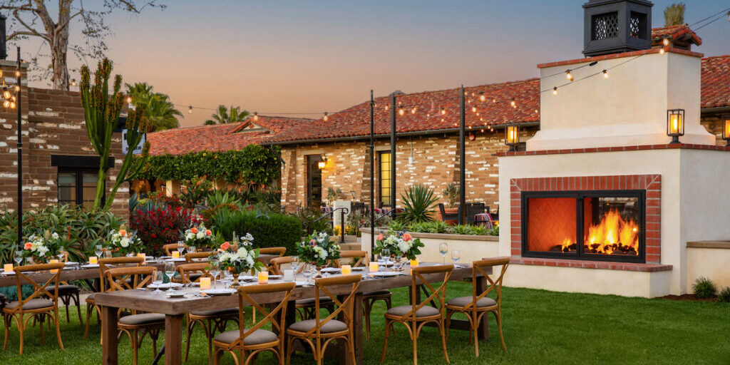 Outdoor dining setup with wooden tables and chairs on a lawn, a lit fireplace nearby, and a brick building in the background at sunset.