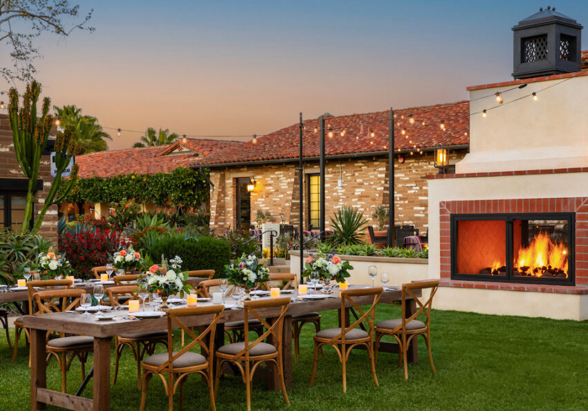 Outdoor dining setup with wooden tables and chairs on a lawn, a lit fireplace nearby, and a brick building in the background at sunset.