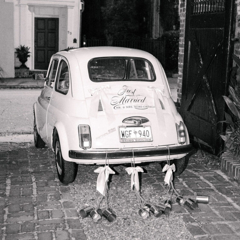A small vintage car decorated with “Just Married” signage and tin cans trailing from the rear bumper.