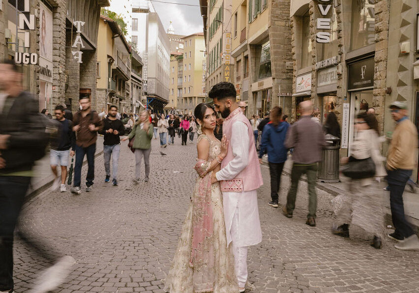 A couple in traditional South Asian attire stands together on a busy cobblestone street, surrounded by blurred pedestrians and historic buildings.