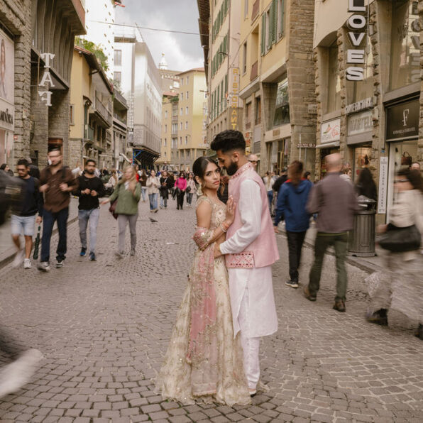 A couple in traditional South Asian attire stands together on a busy cobblestone street, surrounded by blurred pedestrians and historic buildings.