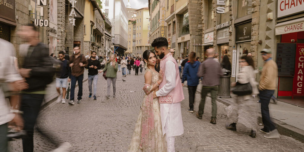 A couple in traditional South Asian attire stands together on a busy cobblestone street, surrounded by blurred pedestrians and historic buildings.