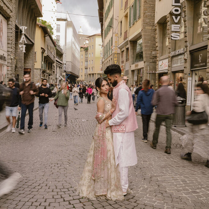 A couple in traditional South Asian attire stands together on a busy cobblestone street, surrounded by blurred pedestrians and historic buildings.