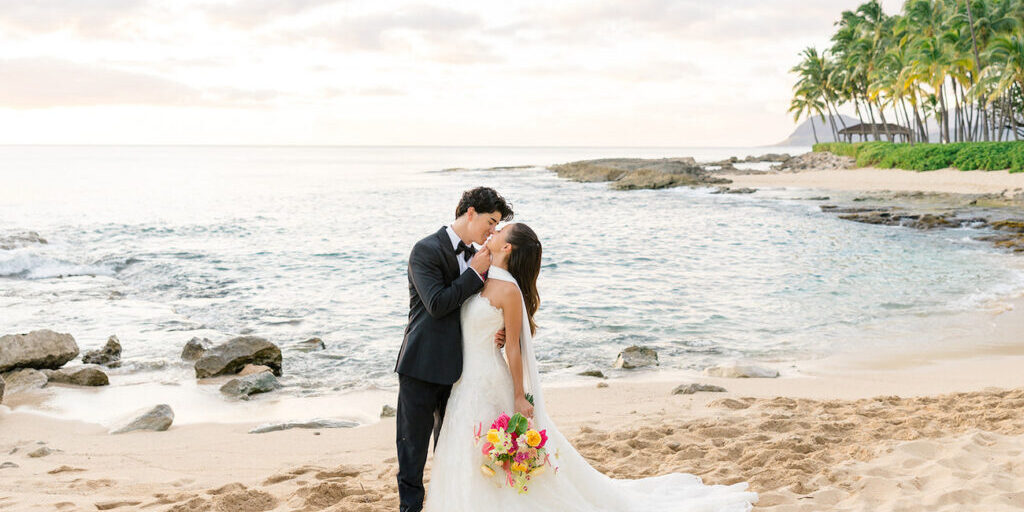 A bride and groom kiss on a sandy beach at sunset, with the ocean and palm trees in the background. The bride holds a colorful bouquet.