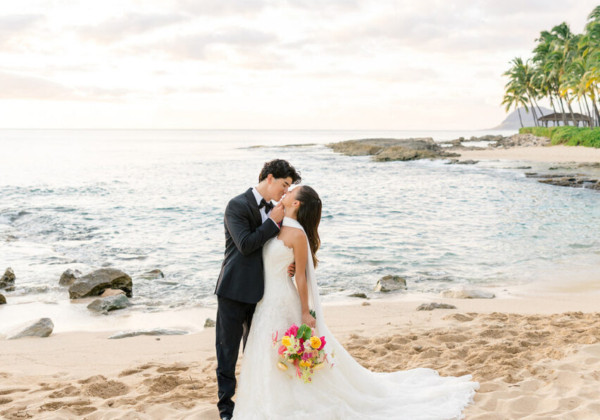 A bride and groom kiss on a sandy beach at sunset, with the ocean and palm trees in the background. The bride holds a colorful bouquet.